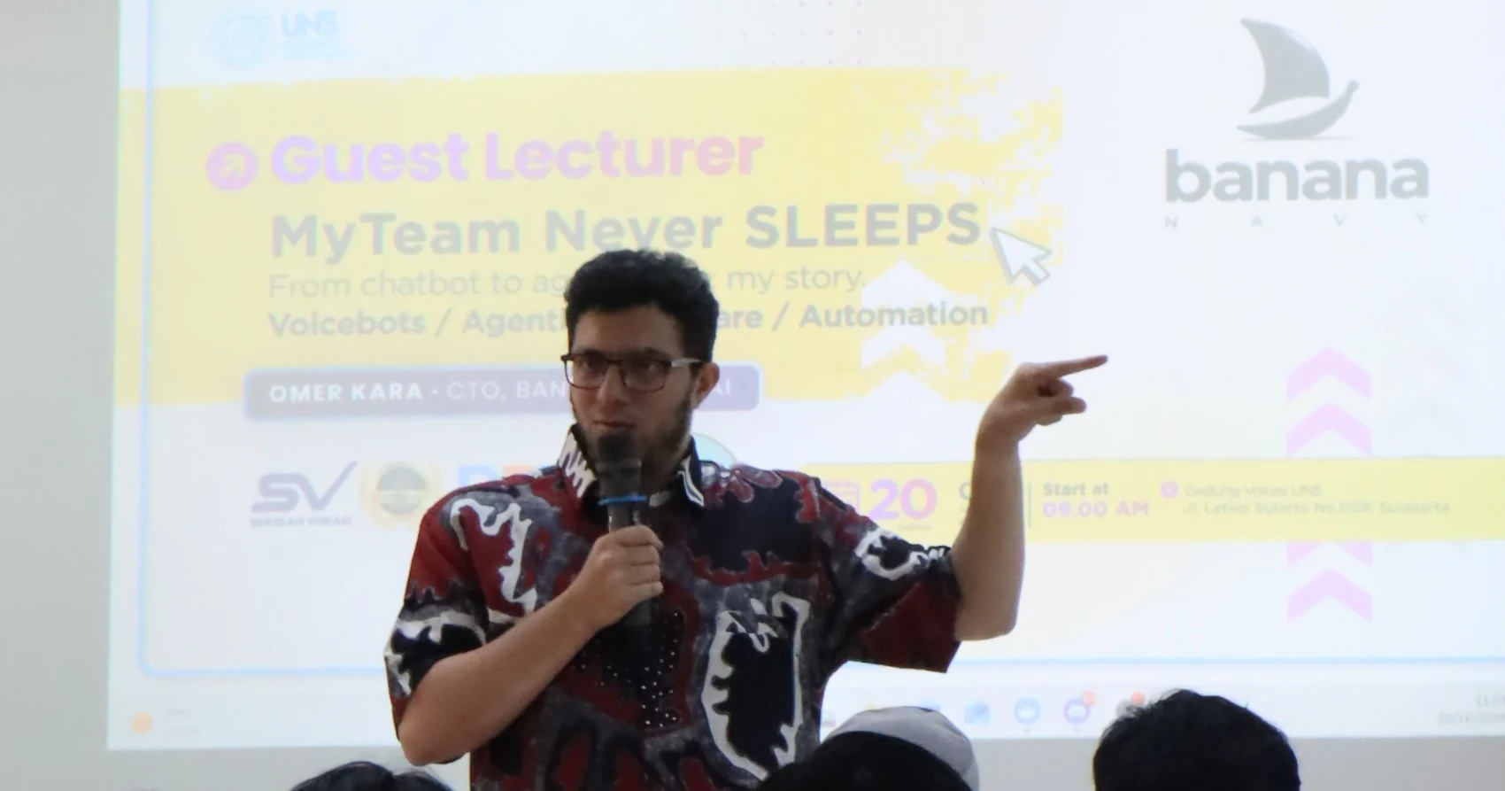 Omer Kara, microphone in hand, points to the side as he speaks, in front of the lecture poster